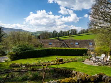 A garden with dining table and chairs near a house with a tiled roof and surrounding trees at The Lodge in Hartlington near Grassington