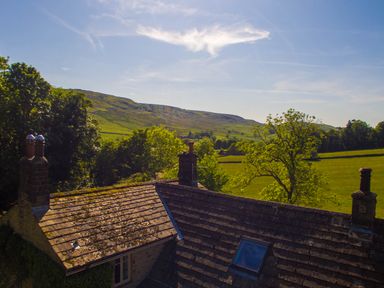 A rooftop with chimneys and skylight set against green trees and hills under a blue sky at The Lodge in Hartlington near Grassington