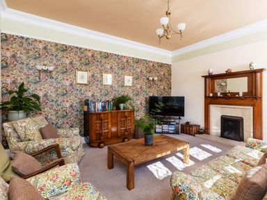 A living room with patterned wallpaper a wooden coffee table floral armchairs a flat screen tv and a fireplace at Kail House in Grassington