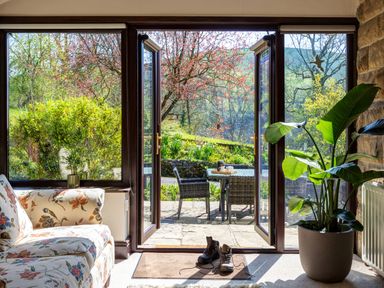 A living room with floral sofa and large potted plant opening to a garden patio with table and chairs at Kail House in Grassington