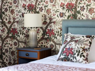A bedroom with a blue headboard floral pillow floral wallpaper and a table lamp on a blue bedside table at Kail House in Grassington