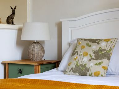 A bedside table with a wicker lamp and rabbit figurine next to a white bed with a pillow printed with rabbits and yellow flowers at Kail House in Grassington