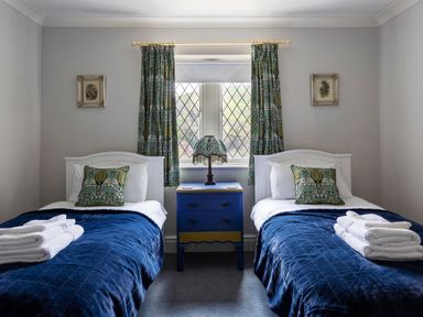 A bedroom with two single beds blue bedspreads green patterned cushions a blue nightstand and a window with patterned curtains at Kail House in Grassington