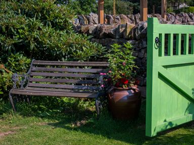 A wooden bench next to a large flower pot with plants beside a green garden gate and a stone wall at How Hill Farm Cottage in Greystoke