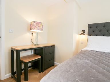 A bedroom corner with a desk, stool, floral lamp, and part of a bed with a grey headboard at How Hill Farm Cottage in Greystoke