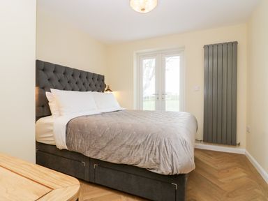 A bedroom with a bed with a tufted headboard and gray bedding near double doors and a vertical radiator at How Hill Farm Cottage in Greystoke