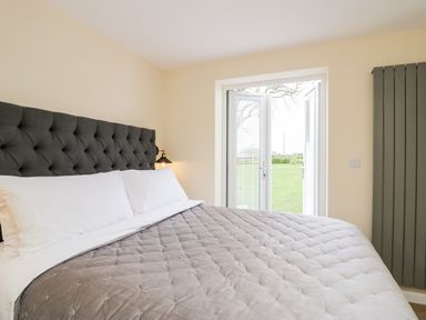 A bedroom with a large bed a grey headboard a wall lamp and glass doors opening to a garden at How Hill Farm Cottage in Greystoke