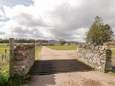A rural farm entrance with a cattle grid and stone walls leading to farm buildings and fields at How Hill Farm Cottage Greystoke