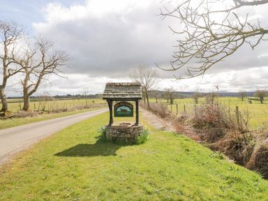 A stone well with a wooden roof and a How Hill Farm sign beside a road with farmland and bare trees at How Hill Farm Cottage in Greystoke