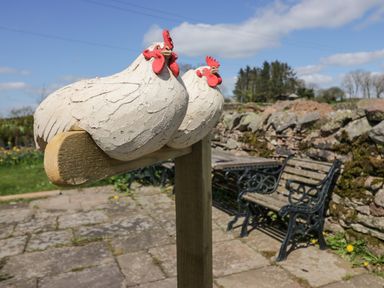 Two decorative chicken sculptures mounted on a wooden post in a garden with stone patio wooden bench and stone wall in the background at How Hill Farm Cottage in Greystoke