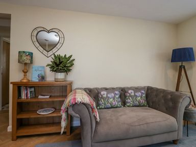 A living room with a brown sofa with floral cushions beside a wooden bookshelf with a lamp and plant at How Hill Farm Cottage in Greystoke