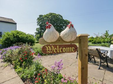 A garden with flowers a wooden welcome sign with two chicken decorations and a bench with a table at How Hill Farm Cottage in Greystoke