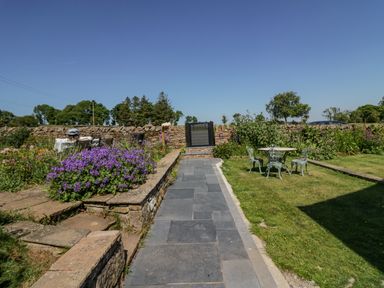 A garden with a stone pathway purple flowers a metal table and chairs and a stone wall at How Hill Farm Cottage Greystoke