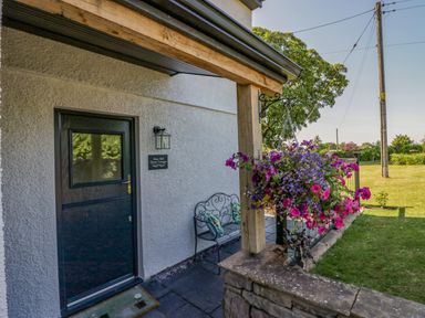 The entrance door of a cottage with a bench with cushions and hanging flowers at How Hill Farm Cottage in Greystoke