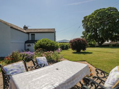 A garden with a table covered with a cloth and chairs with cushions near a white house and trees at How Hill Farm Cottage in Greystoke