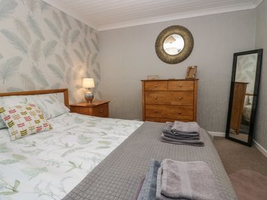 A bedroom with a bed side table lamp wooden chest of drawers and a full length mirror at The Chalet in Coulderton near Egremont Cumbria