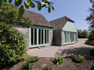 The exterior of a stone building with green framed large windows and doors with a paved pathway and some garden plants at Swallow Barns in Chipping Sodbury