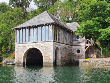A stone and wood boathouse on a body of water with glass windows and surrounding trees at Oakenhurst in Golant