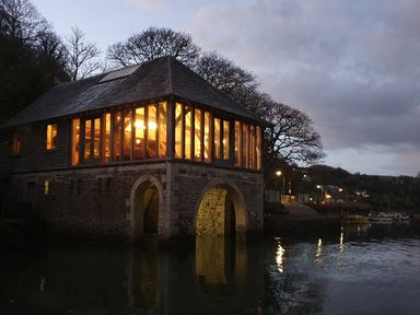 A stone boathouse with large windows and lights on over water at dusk at Oakenhurst in Golant