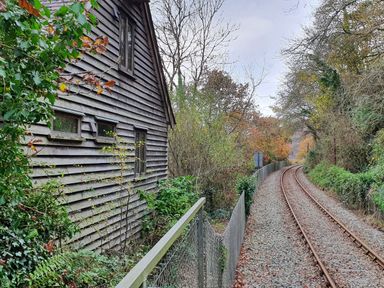 A railway track curving through trees beside a wooden building with small windows and a metal fence alongside the track