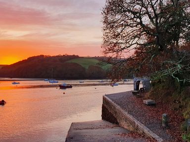 A riverside with boats on the water at sunset and a stone pathway lined with trees at Oakenhurst in Golant