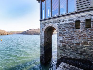 A stone building with an arched opening over water and a river with boats and hills in the background at Oakenhurst in Golant
