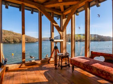 A wooden room with large windows overlooking a body of water with hills in the background and a telescope a small table and a red upholstered bench inside at Oakenhurst in Golant