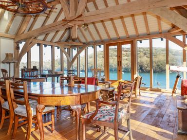 A dining area with wooden table and chairs by large windows overlooking a body of water at Oakenhurst in Golant