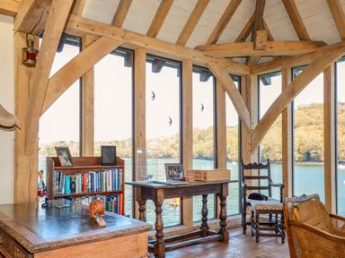 An office with wooden furniture including a desk bookshelf and chairs by large windows overlooking water at Oakenhurst in Golant