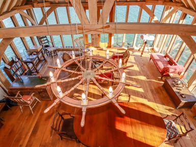A wooden room with large windows overlooking water a round table with chairs a ship wheel chandelier and various seating at Oakenhurst in Golant