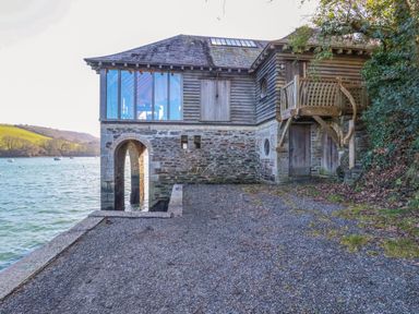 A stone and wood boathouse by the water with a gravel area and trees at Oakenhurst in Golant