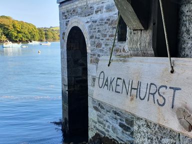 A wooden sign reading Oakenhurst hanging on a stone building by the water with boats and trees in the background at Golant