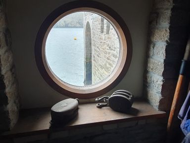 A circular window with a view of a stone arch over water and maritime pulleys on the windowsill at Oakenhurst in Golant
