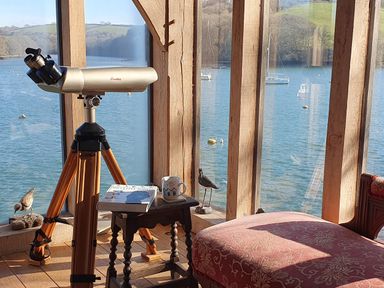 A telescope on a wooden tripod next to a small wooden table with a book and mug by large windows overlooking water with boats at Oakenhurst in Golant