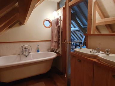 A bathroom with a white bathtub a round window towels on a rack and two sinks with faucets at Oakenhurst in Golant