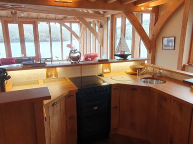 A kitchen with wooden cabinets countertop electric stove and round sink by large windows showing water and hills at Oakenhurst Golant