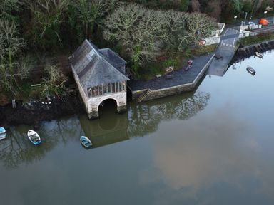A boathouse on a riverbank with small boats in water near a forested area at Oakenhurst in Golant