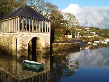 A stone building with arched openings by the water with a small rowboat floating nearby and several boats moored in the background at Oakenhurst in Golant