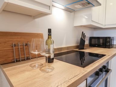 A kitchen countertop with a bottle of wine, two wine glasses, a cutting board, a knife block, stovetop, and microwave at The Bungalow in Lyme Regis