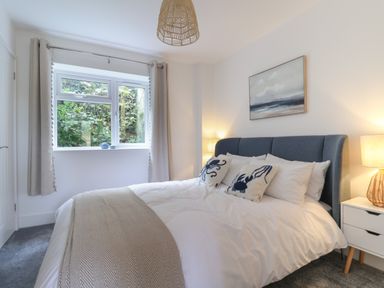 A bedroom with a bed with white bedding and patterned pillows next to a window with curtains and a bedside table with a lamp at The Bungalow in Lyme Regis