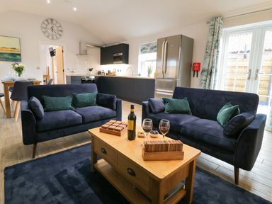 A living room with two navy sofas a wooden coffee table with wine glasses and a bottle and a kitchen with appliances in the background at Bracken Cottage in Pocklington