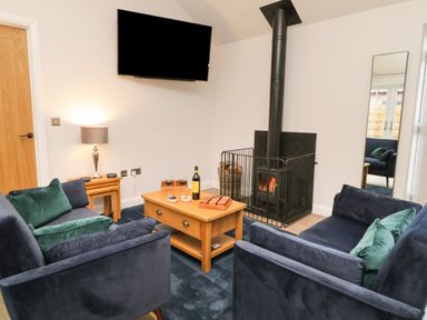 A living room with blue sofas green cushions a wood stove a wooden coffee table with wine and snacks a lamp two nesting tables and a wall-mounted TV at Bracken Cottage in Pocklington