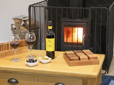A table with two glasses of red wine a bottle of wine bowls of olives a wooden tic tac toe game and a basket of firewood by a fireplace with a fire burning at Bracken Cottage in Pocklington