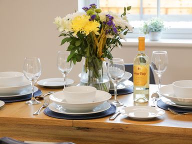 A dining table set with white plates bowls wine glasses cutlery a bottle of white wine and a vase of mixed flowers at Bracken Cottage in Pocklington