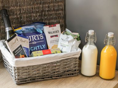 A wicker basket with champagne chocolate crisps biscuits coffee and tea next to bottles of milk and orange juice on a wooden surface at Bracken Cottage in Pocklington