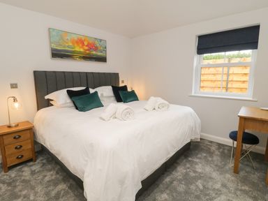 A bedroom with a large bed white bedding and rolled towels on it a wooden nightstand with a lamp a window with a dark shade and a wooden desk with a chair at Bracken Cottage in Pocklington