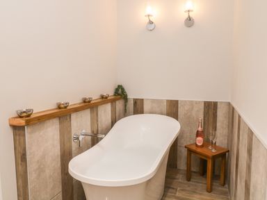 A bathroom with a white freestanding bathtub small wooden table with a bottle and two glasses wall lights and decorative items on a wooden shelf at Bracken Cottage in Pocklington