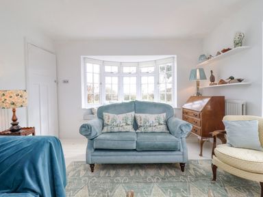 A living room with a blue sofa and cushions a wooden secretary desk and shelves with decorative items at Halsey in Walberswick