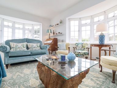 A living room with a blue sofa, armchairs, a glass coffee table, and windows at Halsey in Walberswick