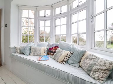 A window seat with multiple cushions and a book on it by large white framed windows at Halsey Walberswick